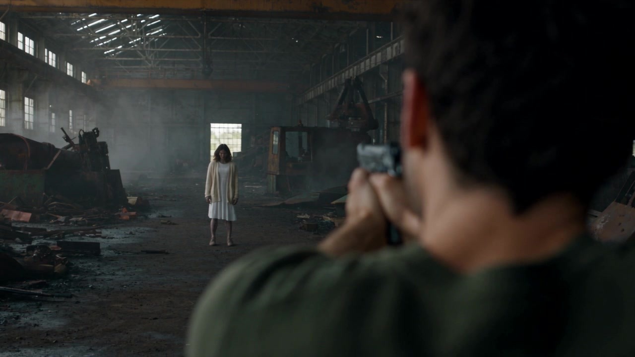 Over-the-shoulder shot of the man aiming a gun at the woman standing far away in a ruined factory