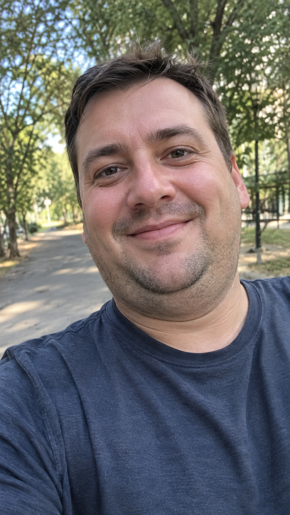 A smiling man in a navy t-shirt takes a close-up selfie outdoors on a tree-lined path, showing a visible double chin.