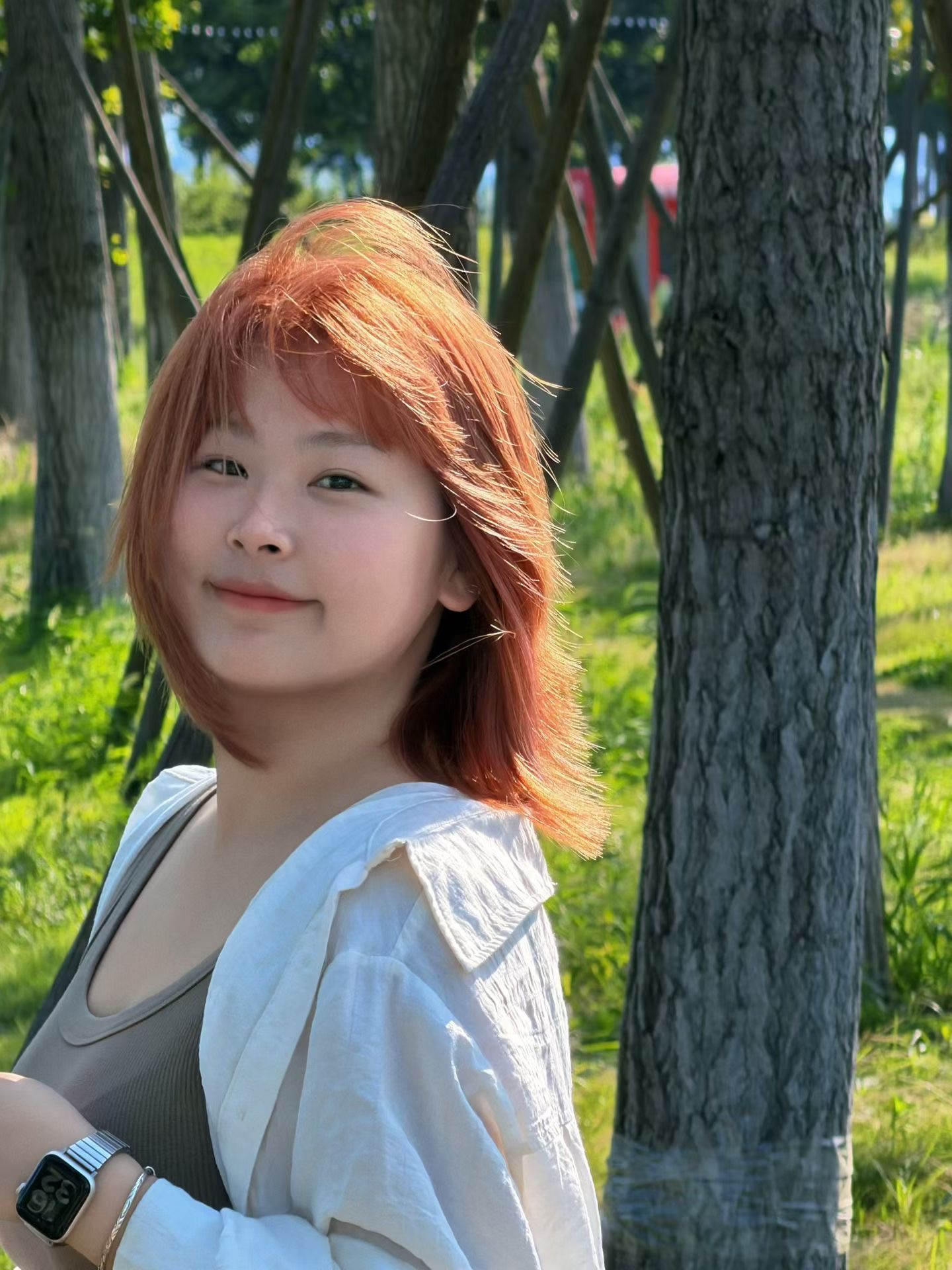 Portrait of a young woman with reddish-brown hair smiling outdoors in a sunlit park with trees.